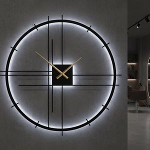May include: A modern, illuminated wall clock with a black frame and gold-colored hands. The clock features a circular design with a glowing white light around the perimeter and a square center. The clock is mounted on a gray wall.
