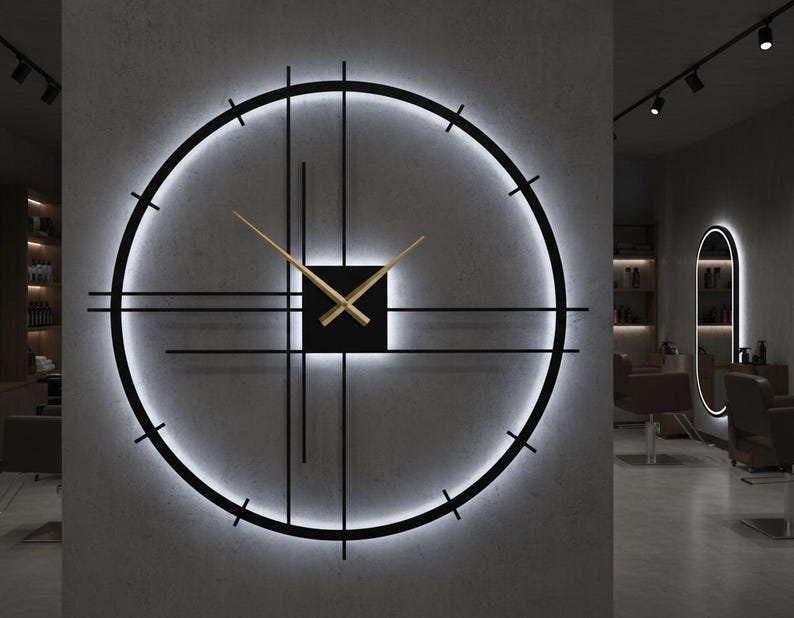 May include: A modern, illuminated wall clock with a black frame and gold-colored hands. The clock features a circular design with a glowing white light around the perimeter and a square center. The clock is mounted on a gray wall.