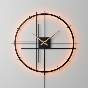 May include: A modern wall clock with a black metal frame and gold-colored hands. The clock features a minimalist design with a circular shape and a warm, orange-colored backlight. The clock's dimensions are not visible.