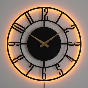 May include: A round, black wall clock with a gold-colored clock hands and Roman numerals. The clock has a glowing orange light around the perimeter. The clock is hanging on a white wall.