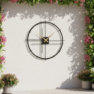 May include: A modern black metal wall clock with gold hands and a minimalist design. The clock is mounted on a white wall, surrounded by lush greenery and pink flowers, creating a stylish outdoor decor element.