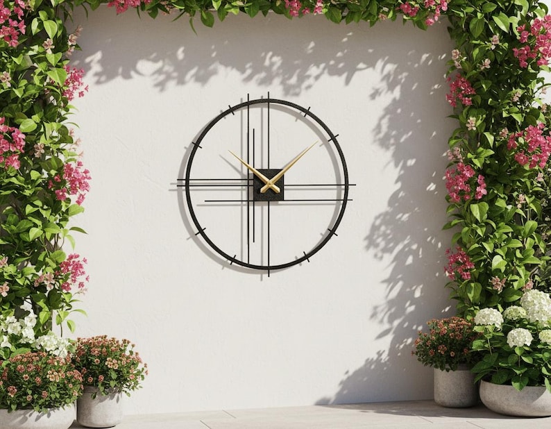 May include: A modern black metal wall clock with gold hands and a minimalist design. The clock is mounted on a white wall, surrounded by lush greenery and pink flowers, creating a stylish outdoor decor element.