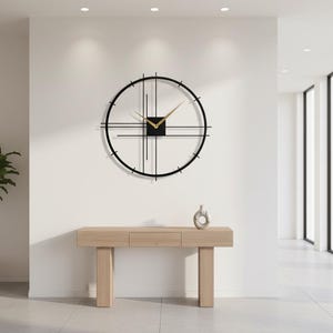 May include: A modern, minimalist wall clock with a black frame and gold hands. The clock is mounted on a white wall above a light wood console table. A small decorative vase sits on the table. A potted plant is to the left.