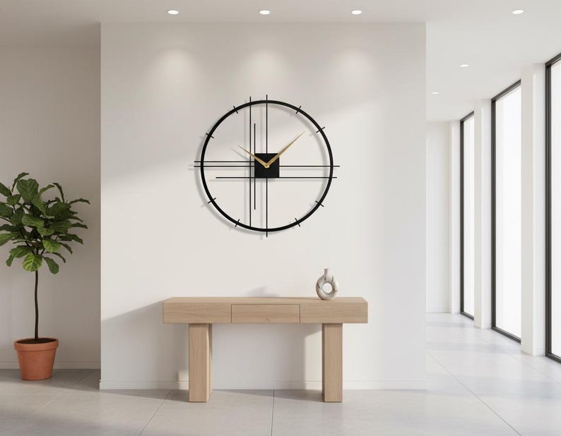May include: A modern, minimalist wall clock with a black frame and gold hands. The clock is mounted on a white wall above a light wood console table. A small decorative vase sits on the table. A potted plant is to the left.