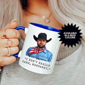 May include: White ceramic mug with a blue rim and handle. The mug features a cartoon illustration of a man wearing a cowboy hat and bandana, with the text "I don't really care, Margaret." The mug is available in 6 colors.