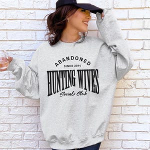 May include: A grey sweatshirt with the text "Abandoned Since 2014 Hunting Wives Social Club" printed on the front.
