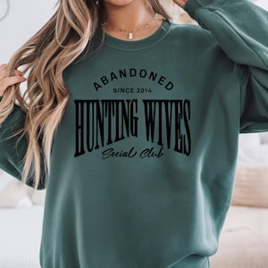 May include: A green sweatshirt with the text "Abandoned Since 2014 Hunting Wives Social Club" in black.