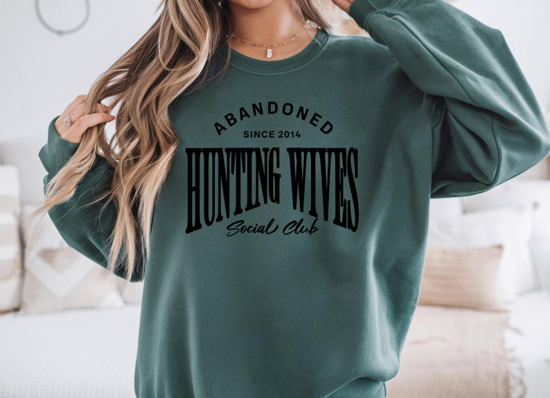May include: A green sweatshirt with the text "Abandoned Since 2014 Hunting Wives Social Club" in black.
