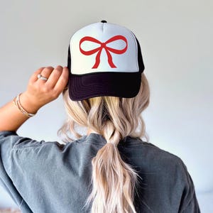 May include: A black and white trucker hat with a red bow on the front. The hat has a mesh back.