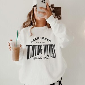 May include: White sweatshirt with black text that reads "Abandoned Since 2014 Hunting Wives Social Club". The person is wearing black and white checked trousers.