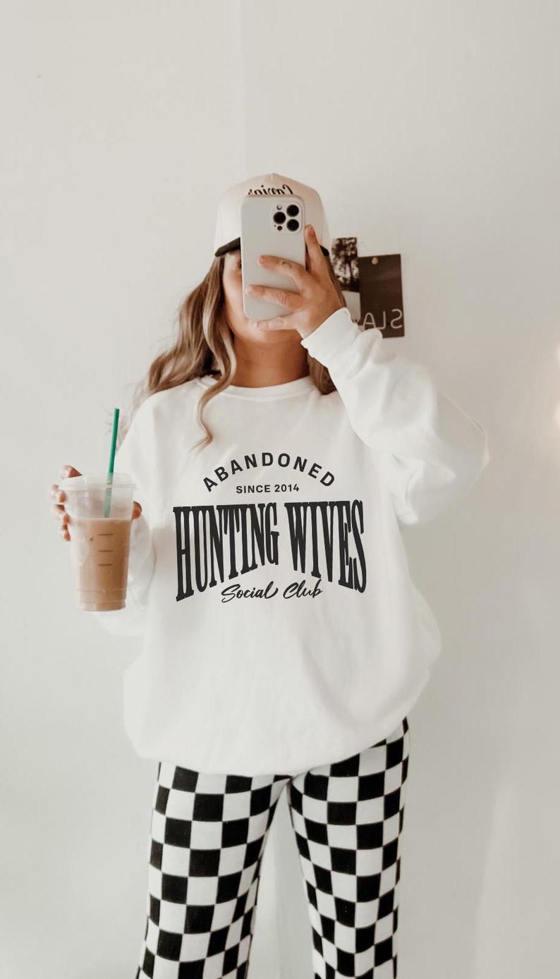 May include: White sweatshirt with black text that reads "Abandoned Since 2014 Hunting Wives Social Club". The person is wearing black and white checked trousers.