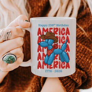 May include: White ceramic mug with a blue balloon dog wearing a cowboy hat and bandana. The mug has the words "Happy 250th Birthday AMERICA" in red and blue, with the years 1776-2026 at the bottom.