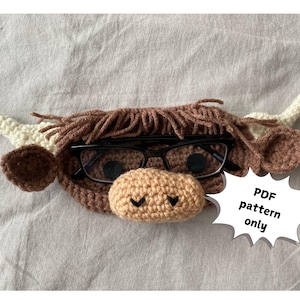 Könnte beinhalten: Ein gehäkeltes Brillenetui in Form einer schottischen Hochlandkuh mit schwarz gerahmten Brillen. Das Etui hat braune, beige und cremefarbene Elemente, mit zotteliger Mähne, Hörnern und Ohren. Der Text "PDF pattern only" steht in einer weißen Sprechblase.
