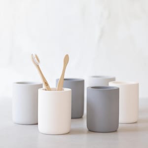 May include: A set of six cylindrical toothbrush holders in various neutral colors: white, light gray, and dark gray. Two bamboo toothbrushes are in one holder. The holders are made of a matte material.
