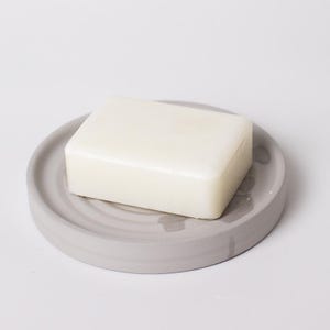 May include: A light gray, circular soap dish with a bar of white soap. The dish has a simple, minimalist design with concentric circles. The soap is rectangular and sits centered on the dish.