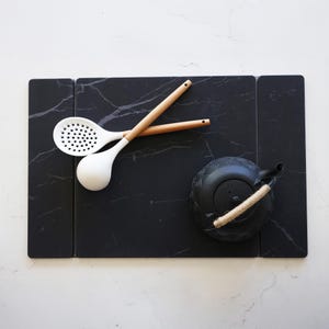 May include: A black marble-look serving board with a white slotted spoon and a white spoon with wooden handles. A black teapot with a rope handle sits on the board. Kitchen and dining decor.