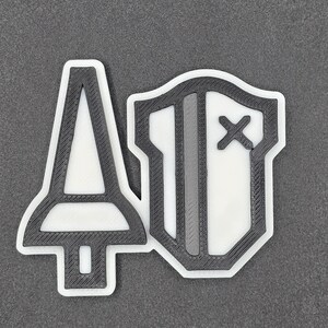 May include: A black and white 3D-printed emblem featuring a stylized sword and shield design. The sword is on the left, and the shield is on the right. The emblem has a white background with black outlines.