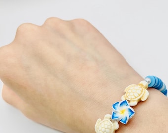 May include: A blue and white beaded bracelet featuring two turtle charms and a blue and white flower charm. The bracelet is worn on a wrist against a white backdrop. The bracelet is made of small, round beads.