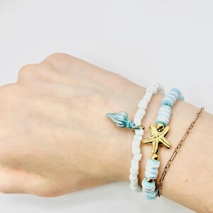 May include: Three beaded bracelets with a beach theme. One bracelet has white beads and a blue seashell charm. Another has white and light blue beads with a gold starfish charm. A gold chain bracelet is also included.
