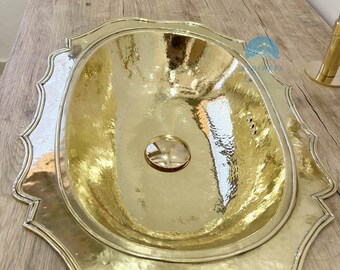 May include: A hammered brass sink with a decorative, scalloped edge. The interior basin is oval-shaped and reflects the light. The sink is set on a light-colored wooden surface, showcasing its metallic sheen.