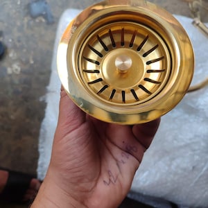 May include: A shiny, gold-colored sink strainer with a circular design. The strainer has a series of vertical slots around the center and a central knob. The product is held in a hand, with the word "BRASS PURE" visible on the side.
