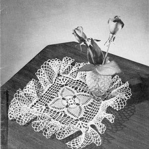 May include: A white, handmade crochet doily with a floral center and scalloped edges. The doily is placed on a dark wooden surface, with a small vase of roses in the center. The image is in black and white.