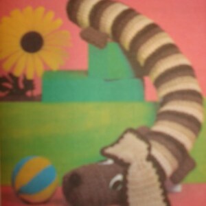 May include: A brown and white striped crocheted dachshund dog toy with a brown head and floppy ears. The toy is sitting on a green and pink surface with a yellow ball.