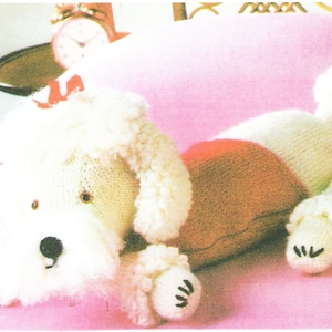 May include: A white knitted poodle toy with a pink and brown knitted sweater. The toy is lying on a pink surface.