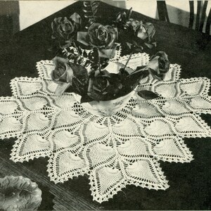 May include: A white crocheted table runner with a scalloped edge and a delicate floral pattern. The runner is centered on a dark wooden table and features a vase of black roses.