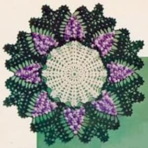 May include: A white crochet doily with a green and purple crochet border. The border features a repeating pattern of leaves and flowers.
