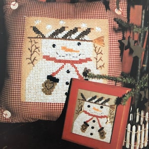 May include: A cross-stitch cushion and framed cross-stitch design featuring a snowman with a black hat, red scarf, and brown mittens. The snowman is surrounded by white snowflakes and black birds.