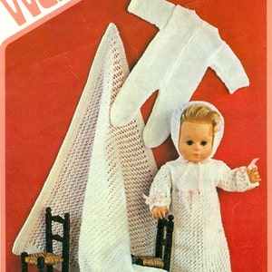 Könnte beinhalten: Ein weißes gestricktes Baby-Puppen-Outfit mit passender Mütze und einer weißen Häkeldecke. Die Puppe sitzt auf einem kleinen Holzstuhl.