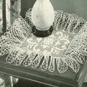 May include: A vintage, white, crocheted doily with a square center and a decorative, scalloped edge. The doily is placed on a dark wooden table, with a lamp on top. The lamp has a round, textured glass shade.