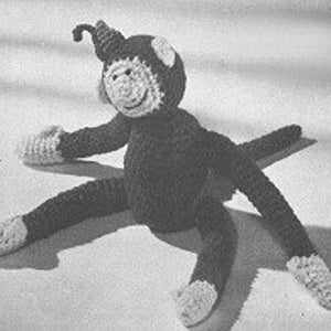 May include: A crocheted monkey toy with a black body, white face, and a long, curled tail. The monkey has four limbs and is sitting on a white surface.