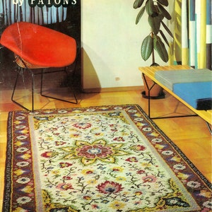 Puede incluir: Una alfombra colorida con un patrón floral en tonos de rojo, amarillo, verde y azul. La alfombra tiene un fondo blanco y un borde marrón oscuro. La alfombra está sobre un suelo de madera en una habitación con una silla roja y una planta.