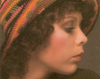 May include: A woman wearing a crocheted hat with stripes of orange, pink, brown, and black. The hat is worn low on her forehead and covers most of her hair. She is wearing a black shirt.