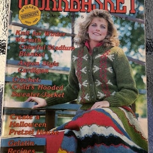 May include: The Workbasket Home Arts magazine cover from October 1985 features a woman wearing a green and white knitted cardigan and a colourful striped blanket. The magazine cover text includes "Knit for Winter Warmth", "Colorful Stadium Blanket", "Aspen Style Cardigan", "Crochet: Child's Hooded Sweater/Jacket", "Create a Halloween Pretzel House", and "Gelatin Recipes".