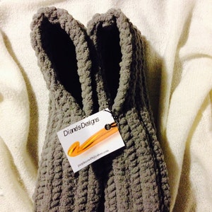 May include: A pair of gray, handmade crochet slippers. The slippers are thick and textured, with a ribbed pattern. A tag attached to the slippers reads "Diane's Designs" and includes an email address. The slippers are resting on a white, textured surface.