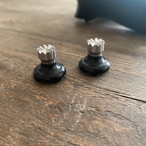 May include: Two black and silver controller thumbsticks on a wooden surface. The thumbsticks have a black base and a silver top with a starburst design. A black controller is visible in the background.