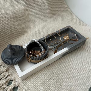 May include: A grey and white jewellery tray with a round, lidded container and a rectangular tray. The container holds a pearl necklace and gold chain. The tray holds gold hoop earrings, a necklace, and spectacles. The items are displayed on a textured beige surface.