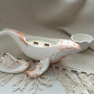 May include: White ceramic whale candle holder with a gold accent. The whale is holding a small wooden wick candle. The candle holder is sitting on a white fringed rug.