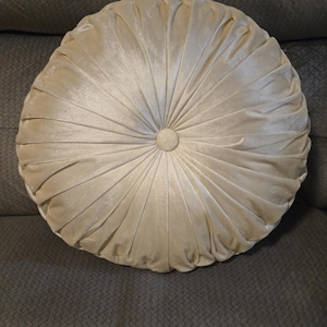 May include: A round, cream-colored velvet pillow featuring a pleated design that radiates from a central, fabric-covered button. The soft, plush fabric creates deep folds and ridges across the surface, giving the cushion a structured, circular appearance.