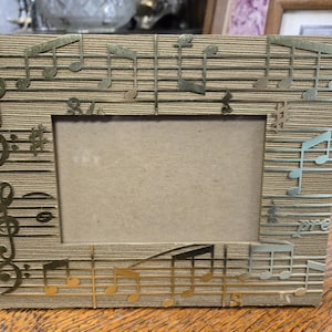 May include: A rectangular photo frame with a light brown, textured background. Gold-colored musical notes and symbols are cut out and applied to the frame. The frame has a rectangular opening for a photo.
