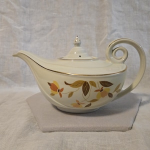 May include: Cream-colored teapot with a curved spout and a looped handle. The teapot features a gold trim and a floral pattern in shades of orange, yellow, and brown. The lid has a small, decorative finial. The teapot is sitting on a gray surface.