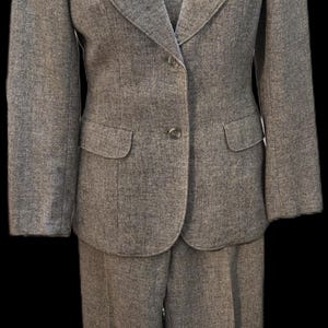 May include: A light gray women's suit, including a blazer, vest, and trousers, displayed on a mannequin. The blazer has two front pockets and a single-button closure. The vest is visible beneath the blazer. The suit is made of a textured fabric.