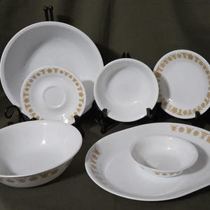 May include: A collection of white ceramic dishware with a gold floral pattern. The set includes various plates, bowls, and a serving platter. The dishes are displayed on black metal stands against a dark green background.