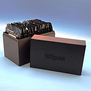 May include: A brown box with a lid, containing individually wrapped black wipes. The lid is black with the word "Wipes" embossed on it. The box is sitting on a light blue surface.
