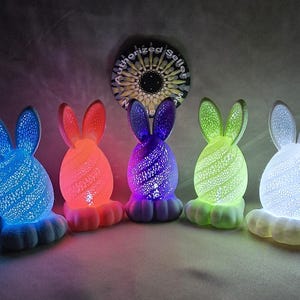 May include: Five ceramic bunny night lights, each with a different coloured light. The bunnies are white with a textured surface and have long ears. The lights are blue, red, purple, green, and white.