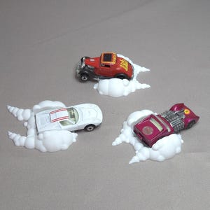 Op de afbeelding: Drie speelgoedauto's, een rode hot rod, een witte sportwagen met rode strepen en een magenta auto, elk gemonteerd op een witte wolk-vormige basis. De rode auto heeft vlammen op de zijkant geschilderd.