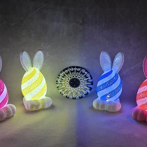Może przedstawiać: Cztery nocne lampki LED w kształcie króliczka wielkanocnego w kształcie jajka. Króliki są białe z różowymi, żółtymi, niebieskimi i czerwonymi paskowanymi jajkami. Lampki są podświetlone.